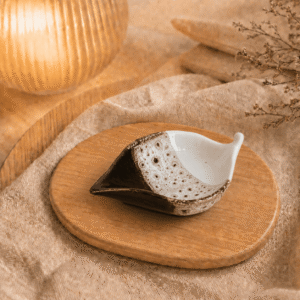 Bruin wit keramisch schaaltje in bladvorm op houten plateau in warme sfeersetting met lamp en beige stof.