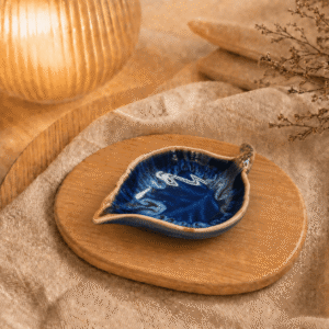 Donkerblauw keramisch schaaltje in bladvorm op houten plateau in warme sfeersetting met lamp en beige stof.