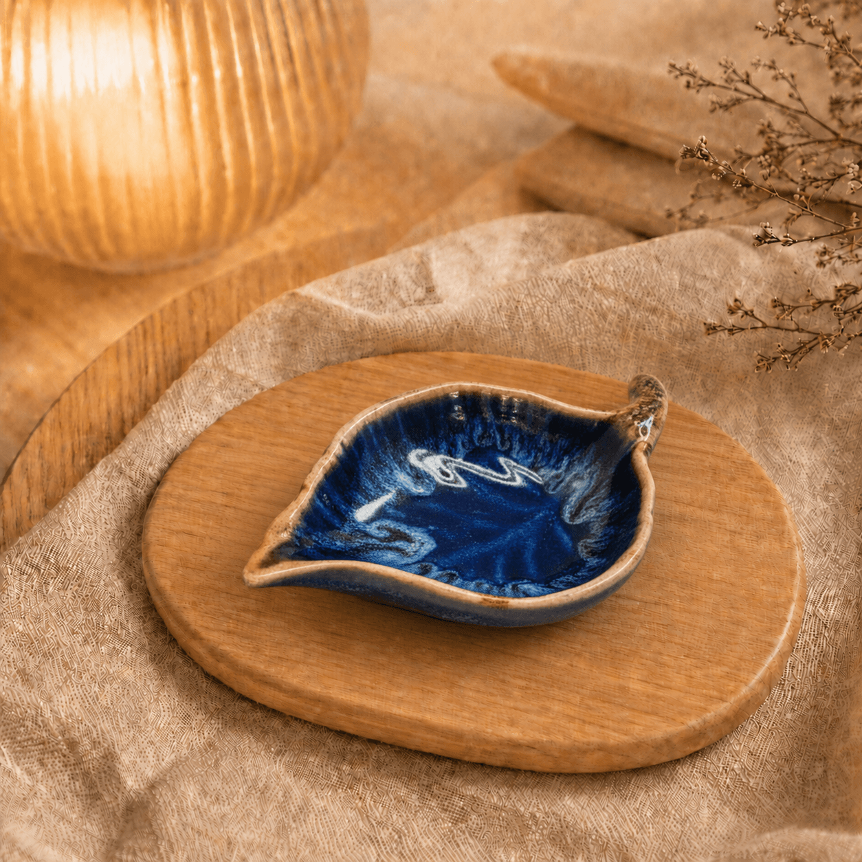 Donkerblauw keramisch schaaltje in bladvorm op houten plateau in warme sfeersetting met lamp en beige stof.