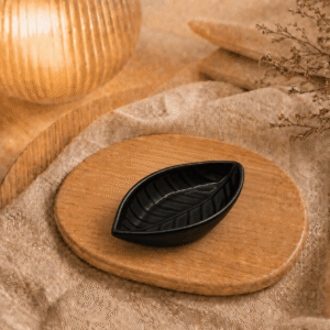 Zwart keramisch schaaltje in bladvorm op houten plateau in warme sfeersetting met lamp en decoratieve takjes.