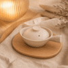 Dekschaal gespikkeld wit met handgreep en deksel op een houten onderzetter met warme verlichting en beige textiel.