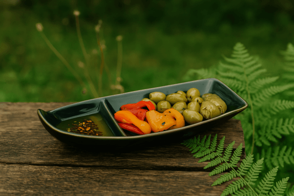 Donkere keramieken bootvormige schaal met twee vakken, gevuld met olijven, gegrilde paprika en dip, buiten gepresenteerd op houten tafel.