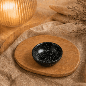 Zwart gespikkeld keramisch schaaltje op houten plateau in warme sfeersetting met zachte verlichting.