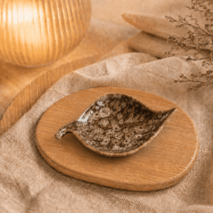 Bladvormig keramisch schaaltje in kastanjebruin met beige spikkelglazuur op houten plateau in warme sfeersetting.