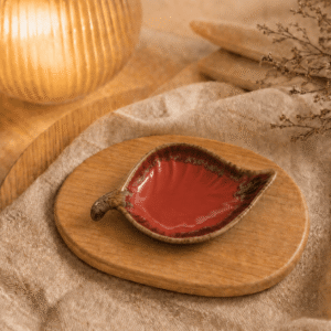 Rood keramisch schaaltje in bladvorm op houten plateau in warme sfeersetting met beige stof, droogbloemen en sfeerlamp.