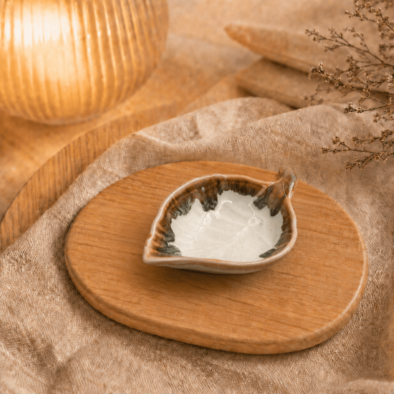 Witbruin keramisch schaaltje in bladvorm op houten plateau in warme sfeersetting met lamp en beige stof.