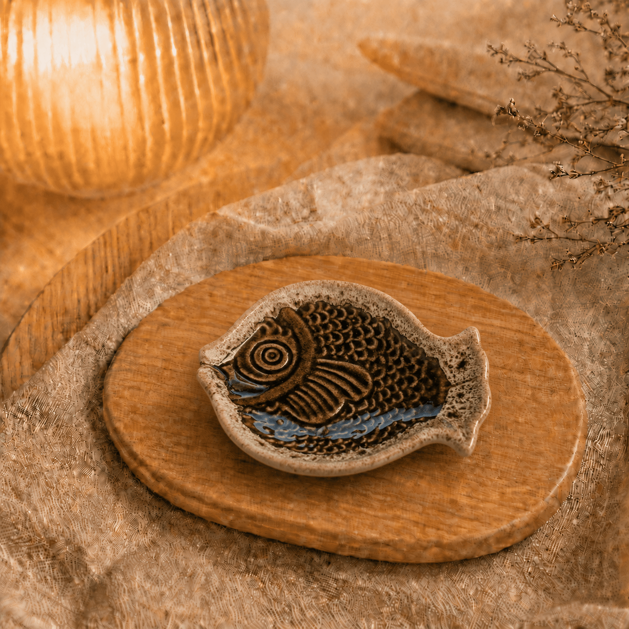 Bruin keramisch visschaaltje op houten plateau in warme sfeersetting met lamp en decoratieve takjes.