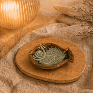 Lichtgroen keramisch visschaaltje op houten plateau in warme sfeersetting met lamp en beige stof.