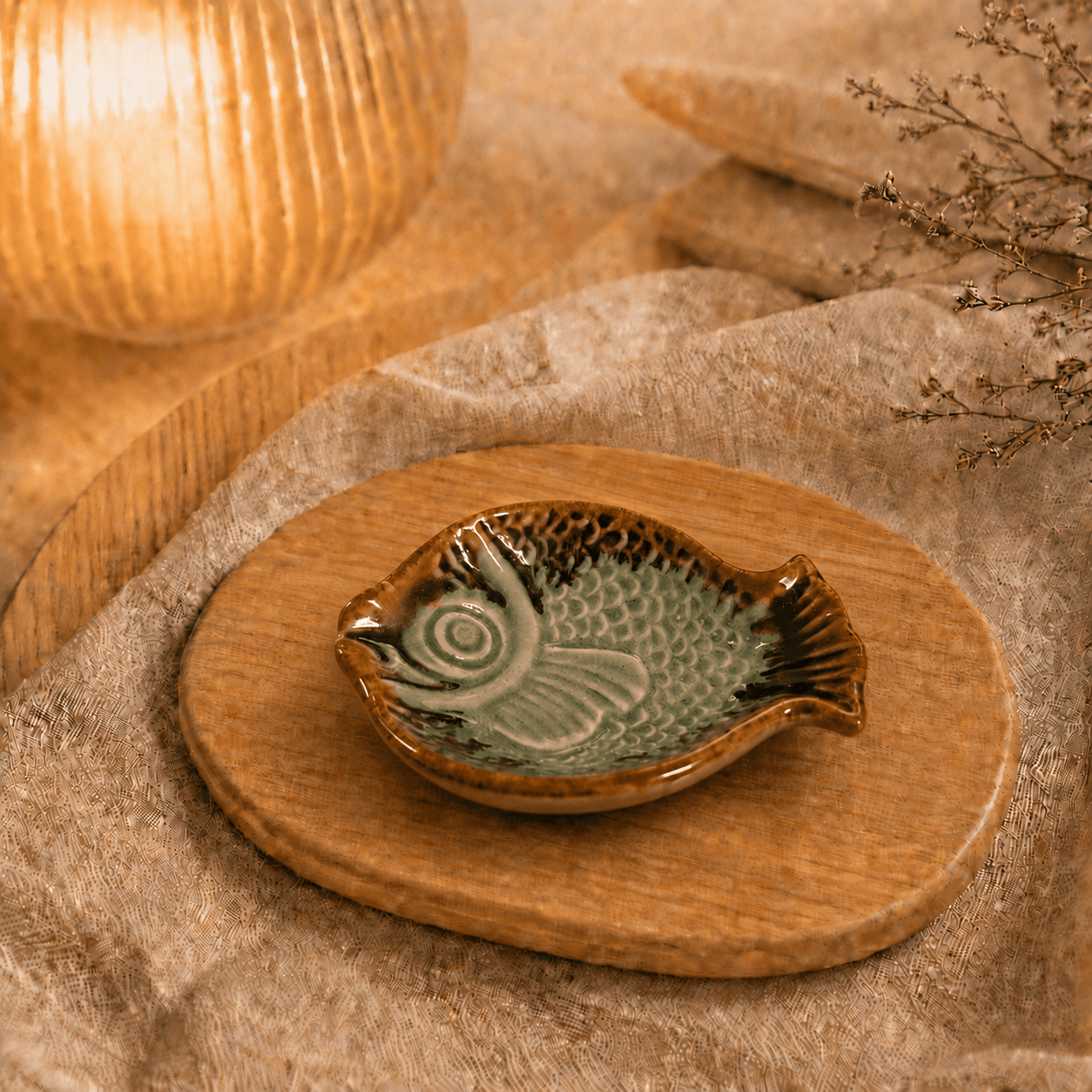 Lichtgroen keramisch visschaaltje op houten plateau in warme sfeersetting met lamp en beige stof.