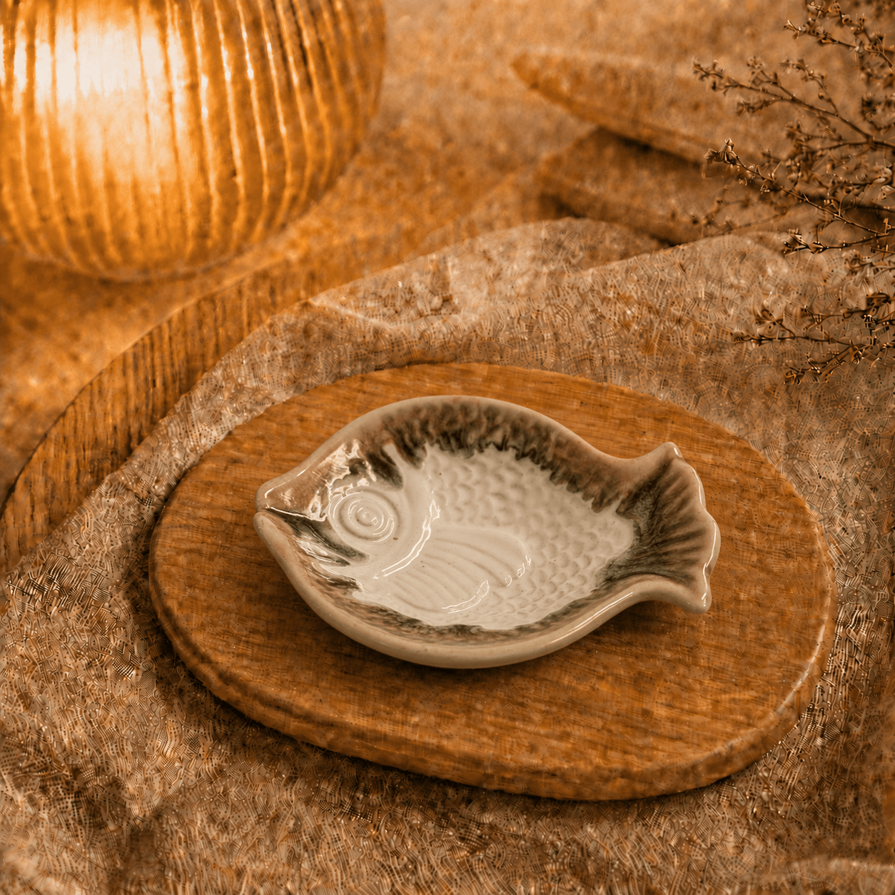 Wit keramisch visschaaltje met bruine accenten op houten plateau in warme sfeersetting met lamp en decoratieve takjes.
