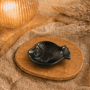 Zwart keramisch visschaaltje op houten plateau in warme sfeersetting met lamp en decoratieve takjes.
