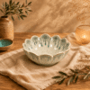Grijs-groene keramieken lotuskom van 13 cm op een beige linnen doek en houten tafel, sfeervol gecombineerd met olijftakken, een aardewerken vaas en warm kaarslicht.
